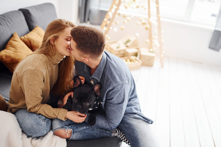Happy young modern couple sitting on the sofa at home with christmas tree with their cute dog.