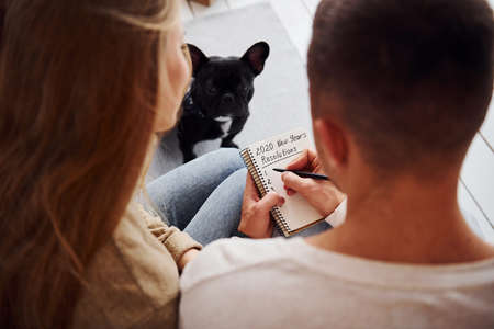 Happy young modern couple sitting on the sofa and writing wishes on notepad at home with their cute dog.の写真素材