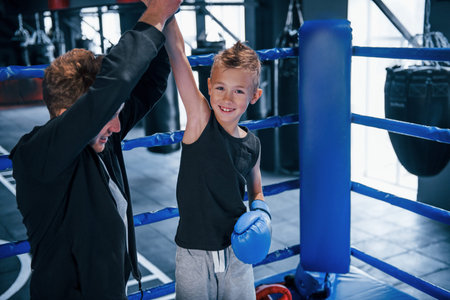 Boxing coach standing in the ring with boy and celebrating victory together.