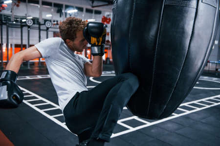 Young man in white shirt and boxing protective gloves doing exercises in gym with pushing bag.の写真素材