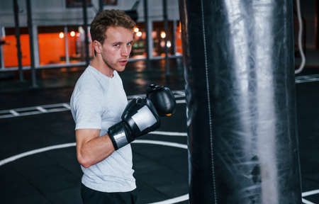 Young man in white shirt and boxing protective gloves doing exercises in gym with pushing bag.の写真素材