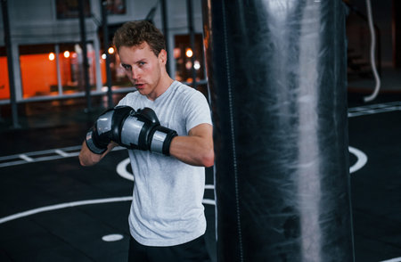 Young man in white shirt and boxing protective gloves doing exercises in gym with pushing bag.