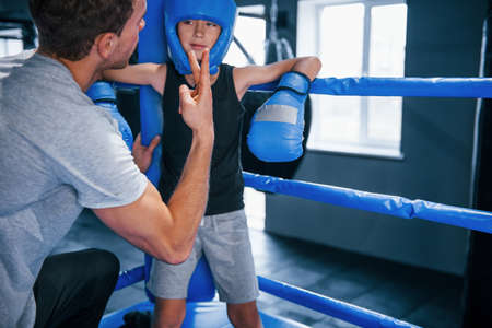 Young boxing coach is helping little boy in protective wear on the ring between the rounds.の写真素材