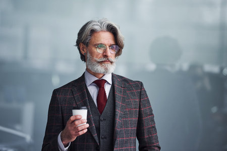 Senior businessman in suit and tie with gray hair and beard standing with cup in hand.