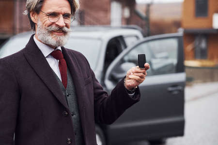 Fashionable senior man with gray hair and beard standing outdoors on the street near his car with keys in hand.の写真素材
