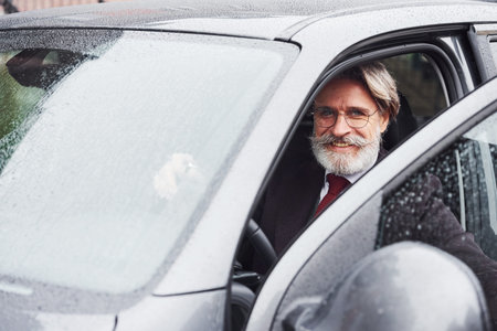 Fashionable senior man with gray hair and beard is outdoors on the street sitting in his car.