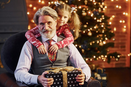 Cheerful fashioned senior man with grey hair and beard with little girl with gift box embracing each...
