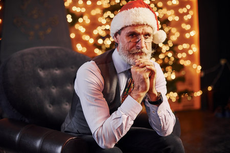 Portrait of stylish senior with grey hair and beard sitting in decorated room and in christmas hat.