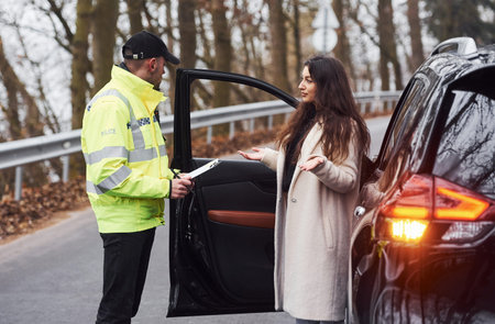 Male police officer in green uniform talking with female owner of the car on the road.