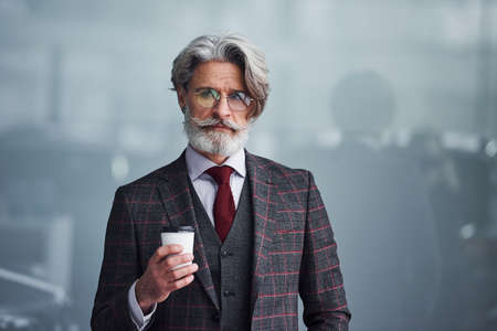 Senior businessman in suit and tie with gray hair and beard standing with cup in hand.の写真素材