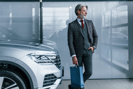 Senior businessman in suit and tie with gray hair and beard standing indoors with blue package near...