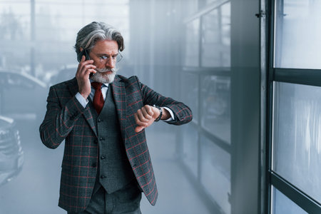 Senior businessman in suit and tie with gray hair and beard standing indoors with phone looking at...