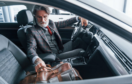Fashionable senior man with grey hair and mustache on vacation with bag inside modern automobile.