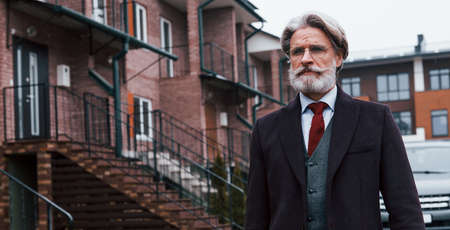 Fashionable senior man with gray hair and beard walks outdoors on the street against his car.の写真素材