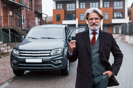 Fashionable senior man with gray hair and beard standing outdoors on the street near his car with...