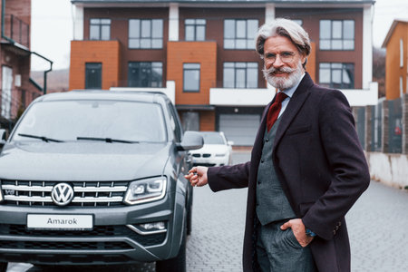 Fashionable senior man with gray hair and beard standing outdoors on the street near his car with...