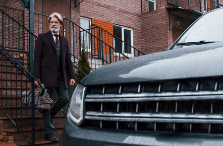 Fashionable senior man with gray hair and beard walks with bag outdoors on the street near his car.
