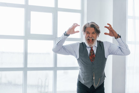 Screaming senior man in jacket and tie indoors in room against window.