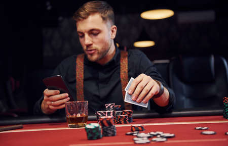 Elegant young man sits in casino, using phone and plays poker game.の写真素材