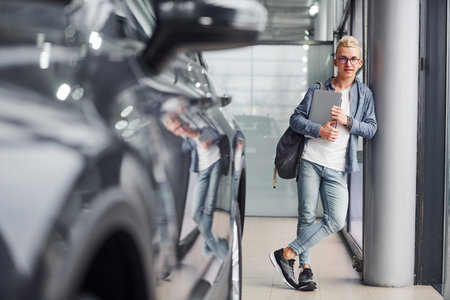 Young hipster guy in nice clothes stands indoors against grey background and near car.