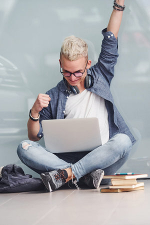 Young hipster guy sitting indoors near grey background with laptop and books.