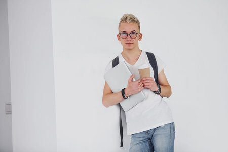 Student in casual clothes and with backpack stands indoors against white wall with cup of drink and...