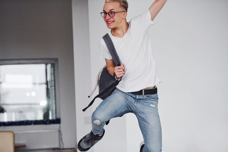 Happy hipster guy in casual clothes and with backpack jumping indoors against white wall.
