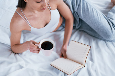 Positive smiling woman in sportive clothes lying down in the studio with book and cup of drink in...