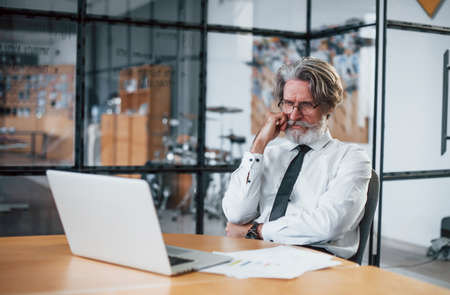 Bored mature businessman with grey hair and beard in formal clothes is in the office against laptop.の写真素材