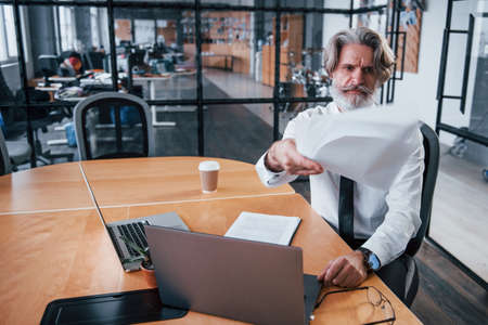Throwing documents. Mature businessman with grey hair and beard in formal clothes is in the office.の写真素材