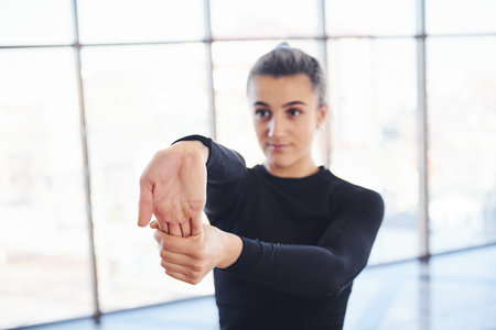 Confident young sporty woman in sportswear doing warm up exercises for hands against window.