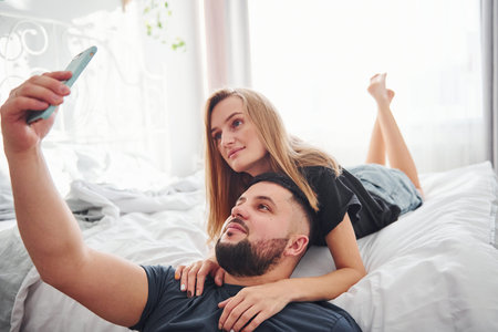 Young married couple making a selfie by using phone in bedroom at daytime.