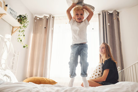 Mother playing pillow fight with her son in bedroom at daytime.