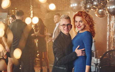 Young couple standing together against their friends in christmas decorated room and celebrating New...