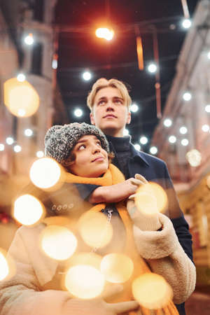 Positive young couple in warm clothes embracing each other on christmas decorated street.の写真素材