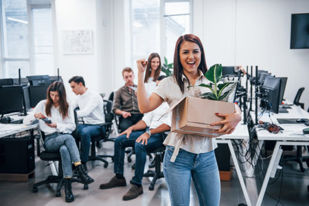 Woman smiling and holding box with green plant inside. Standing against people that working in...