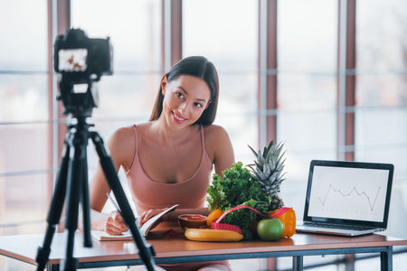 Young fitness vlogger doing video indoors by sitting near table with healthy food.