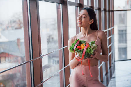 Girl in sportive wear and with healthy food in hands stands indoors near window.の写真素材