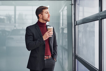 Young business man in luxury suit and formal clothes wth cup of drink is indoors in the office.