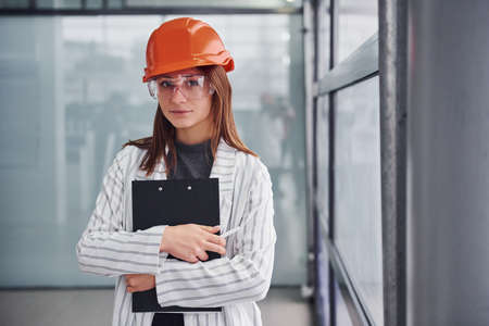 Portrait of young female worker with protective helmet, eyewear and notepad. Standing in the office.の写真素材