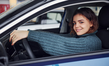 Positive young girl in casual clothes sitting inside of modern car and smiling.