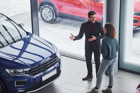 Professional salesman assisting young girl by choosing new modern automobile indoors.