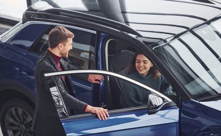 Professional salesman assisting young girl by choosing new modern automobile indoors.
