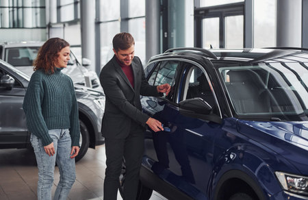 Professional salesman assisting young girl by choosing new modern automobile indoors.