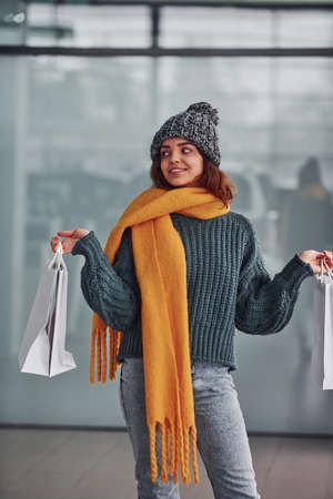 Smiling and feeling happy. Beautiful cheerful girl in yellow scarf and in warm clothes standing indoors with shopping bags in hands.の写真素材