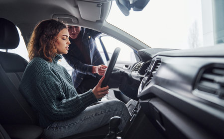 Professional salesman assisting young girl by choosing new modern automobile indoors. Woman sitting...