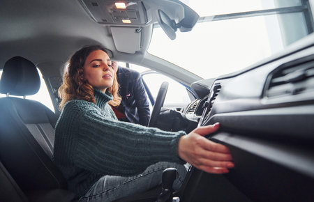 Professional salesman assisting young girl by choosing new modern automobile indoors. Woman sitting...