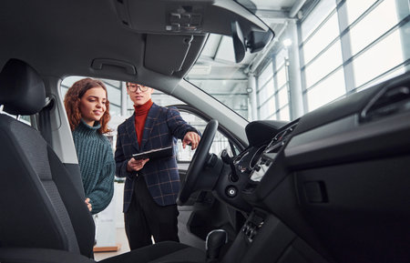 Professional salesman assisting young girl by choosing new modern automobile indoors. Woman sitting...