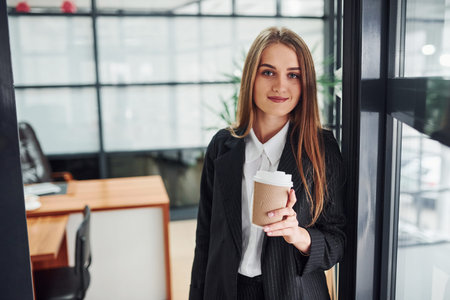 Woman in formal clothes standing indoors in the office with cup of drink.の写真素材