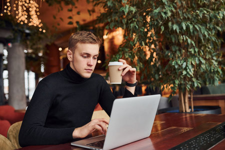 Successful young guy sits in cafe by table with his laptop on it.